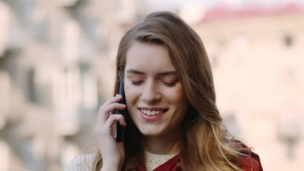 Hymyilevä tyttö puhuu puhelimessa kadulla. Onnellinen nainen käyttää puhelinta ulkona — kuvapankkivalokuva