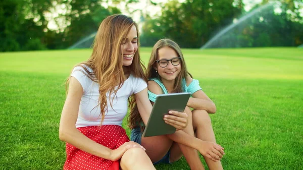 Äiti ja tytär nauravat puistossa. Nainen ja tyttö puhuvat kameran tabletilla — kuvapankkivalokuva