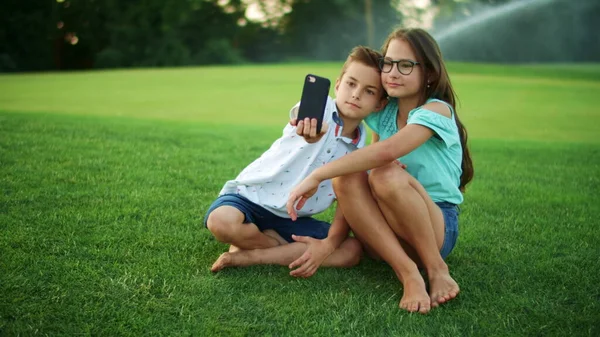 Átöleli a testvérét a parkban. Mosolygós fiú szelfit csinál a mobilján a lánnyal. — Stock Fotó