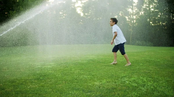 Lykkelig gutt som leker med vannsprederen i åkeren. Positiv fyr hopper i luften – stockfoto