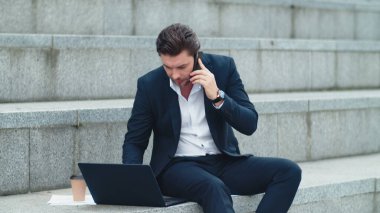 Sokakta dizüstü bilgisayarda çalışan bir iş adamı. Yönetici akıllı telefondan konuşuyor.