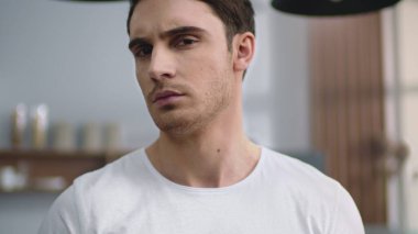 Ev mutfağında kameraya bakan ciddi bir adam var. Odaklanmış adam el ele tutuşuyor.