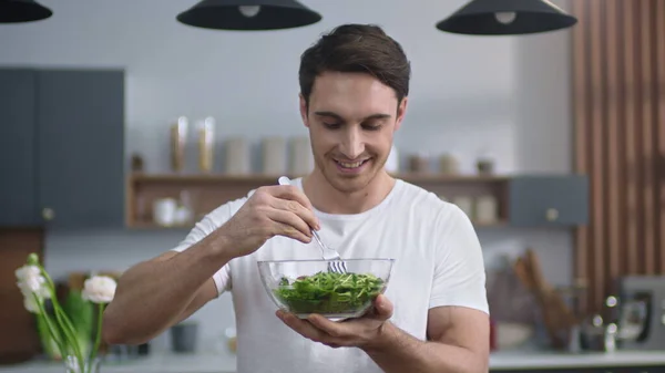 Hymyilevä mies syö tuoretta salaattia kotikeittiössä. Urheilullinen kaveri nauttii terveellistä ruokaa — kuvapankkivalokuva