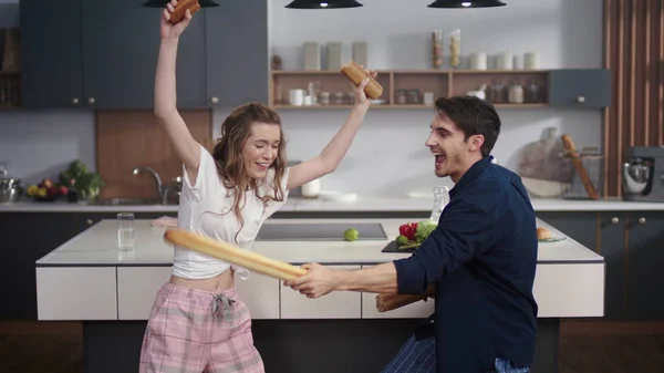 Pareja peleando con baguette en la cocina casera. Amigos alegres divirtiéndose en casa — Foto de Stock