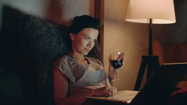 Flicka surfa på internet på bärbar dator på soffan. Kvinna dricker rött vin med laptop. — Stockfoto