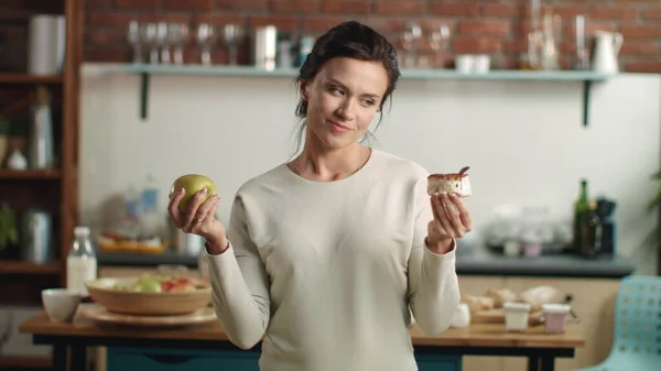 Nainen tekee valinnan omenan ja kakun välillä keittiössä. Tyttö valitsemalla hedelmiä ruokavalio. — kuvapankkivalokuva