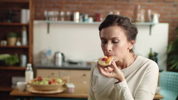 Nainen katselee kameraa hieno kakku kädessään. Tyttö puree jälkiruokaa kotona. — kuvapankkivalokuva