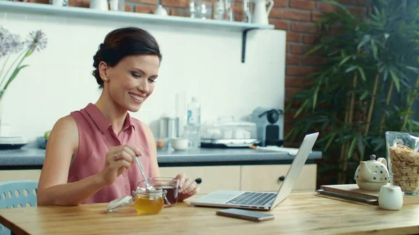Femme utilisant un ordinateur portable pour un appel vidéo en ligne. Fille verser du miel dans une tasse de thé. — Photo
