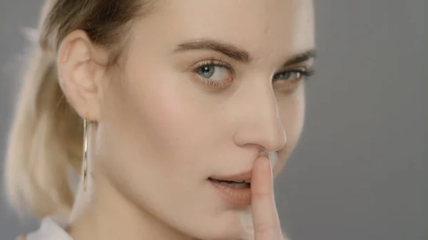 Young woman showing silence sign. Blonde girl putting finger on mouth in studio