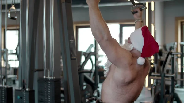 Seksi Noel Baba spor salonunda göğüs presi yapıyor. Spor kulübünde Noel Baba şapkasıyla formda adam eğitimi. — Stok fotoğraf