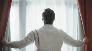 Arka planda perdeleri açan genç adam. Yakından bakan yakışıklı adam pencerenin yanında duruyor..