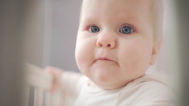 Mavi gözlü şirin bebek yüzlü. Bebek yatakta dikilip etrafa bakıyor.