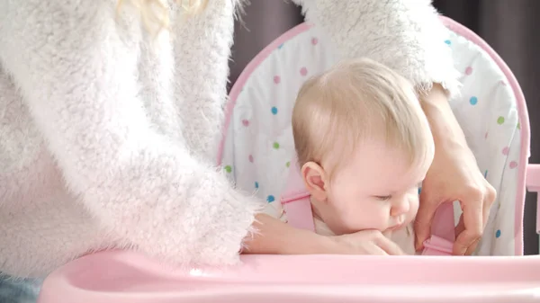 Mother putting baby in chair in kitchen. Mom hands fasten belts in pink chair — Stock Photo, Image