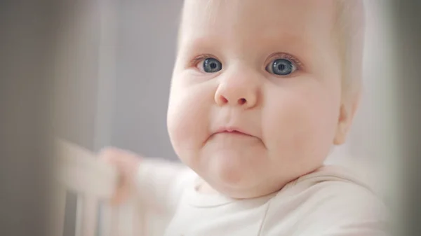 Linda cara de bebé con ojos azules. Bebé de pie en la cama y mirando a su alrededor —  Fotos de Stock
