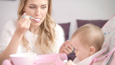 Sarışın kadın çocuğu kaşıkla besliyor. Annem bebeği püre yiyecekle besliyor.