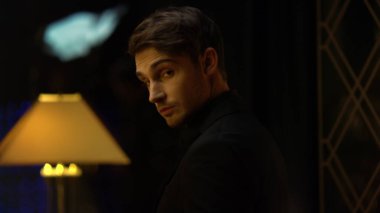 Karanlık odada şık bir adam duruyor. Zarif bir adam kamerayı içeri çeviriyor..