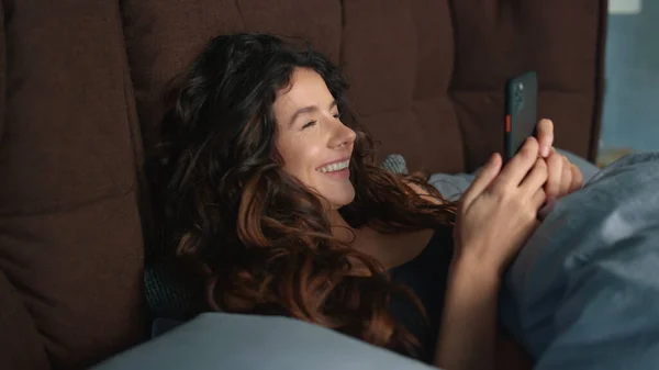 ベッドで携帯電話を使って幸せな女性。陽気な女の子見る携帯電話の画面. — ストック写真