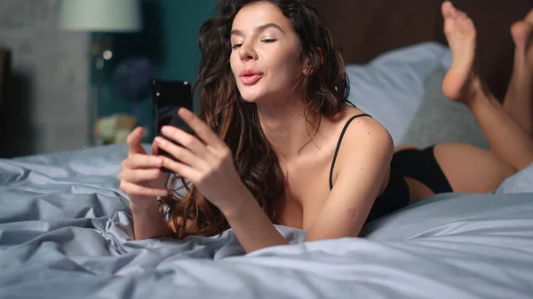 陽気な女性は電話ベッドをリラックス。魅力的な女の子ビデオ通話スマートフォンを持っている — ストック写真