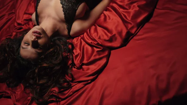 Ragazza sexy in cerca di fotocamera sul letto di seta rossa. Affascinante donna rilassante lenzuola di raso. — Foto Stock