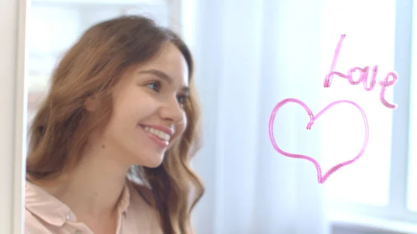 Young woman writing by lipstick word love on mirror with heart in bathroom