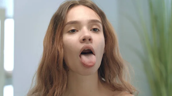 Young woman with opened mouth looking tongue and throat in mirror in bathroom