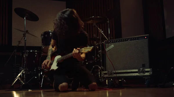 Expressive guitarist playing electric guitar on knees in recording studio.