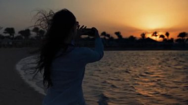 Deniz kenarında cep telefonuyla günbatımının fotoğrafını çeken çekici bir kadın..