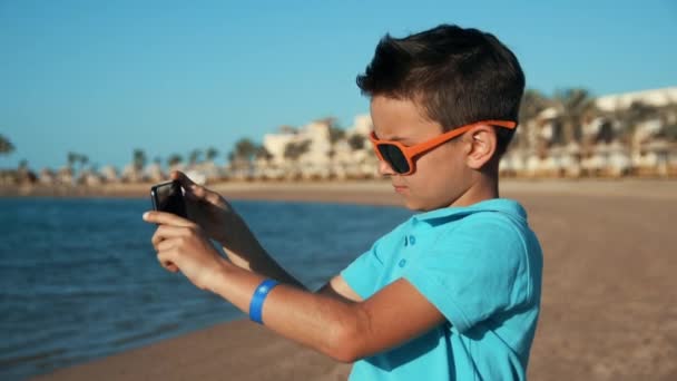 Привабливий хлопчик-підліток фотографує мобільний телефон на сонячній береговій лінії . — стокове відео