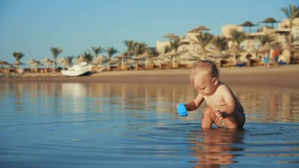 ぽっちゃり赤ちゃんは海水でおもちゃで遊んでいます。海辺で子供が水を撒く — ストック動画