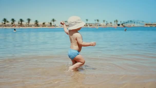 Menino doce dando os primeiros passos na água do mar. Bebê bonito espirrando água — Vídeo de Stock