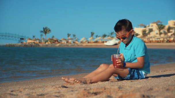Aantrekkelijke tiener met een zonnebril die limonade drinkt op het strand. Jongen zet glas — Stockvideo