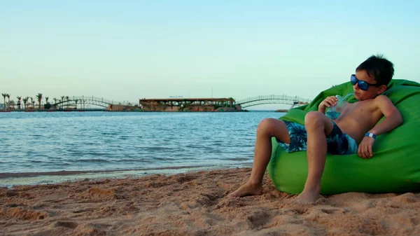 Junger Mann trinkt Saft an der Küste. Teenager sitzt in Stuhlsack am Strand. — Stockfoto