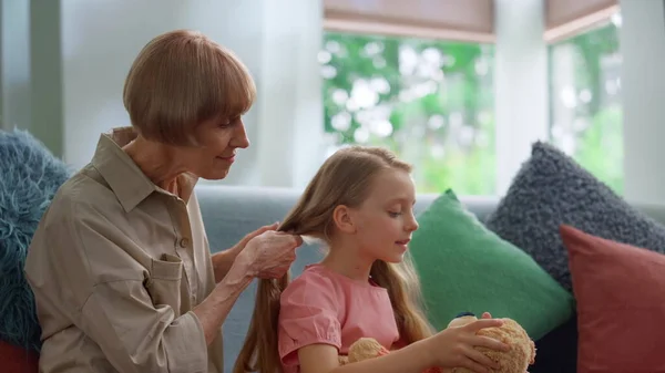 Radosna babcia warkocze włosy małej wnuczki w salonie. — Zdjęcie stockowe