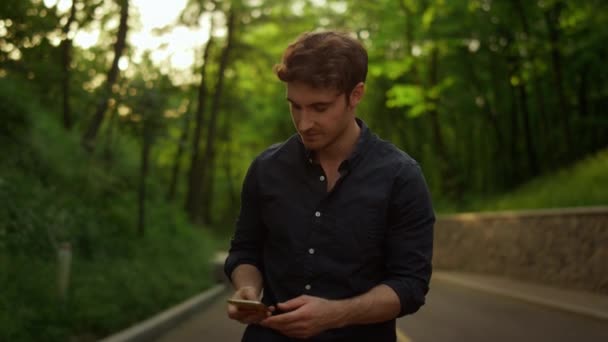 Un hombre guapo sacando el teléfono del bolsillo al aire libre. Guy mensaje de texto en el parque — Vídeos de Stock