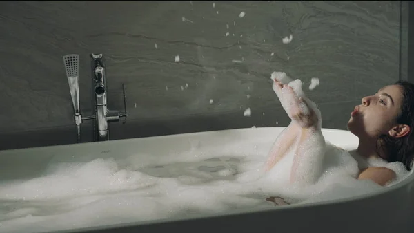 Chica alegre soplando espuma en la bañera. Mujer sonriente tocando la piel en el baño. — Foto de Stock