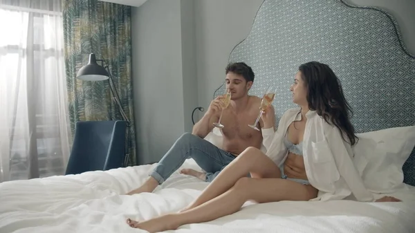 Linda pareja tostando vasos en el apartamento. Encantadora pareja sentada en la cama del hotel. —  Fotos de Stock