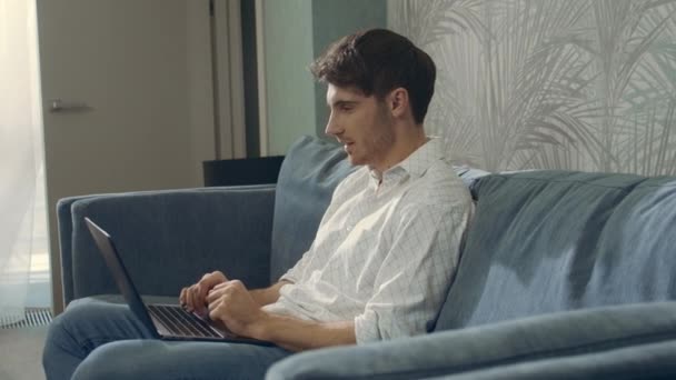 Un uomo che ride leggendo il computer dei messaggi. Felice ragazzo seduto divano con computer portatile. — Video Stock