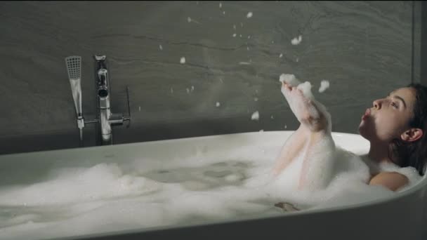 Wesoła dziewczyna dmuchająca pianę w wannie. Uśmiechnięta kobieta dotykająca skóry w wannie. — Wideo stockowe