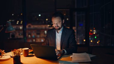 Gece panoramik dairede dizüstü bilgisayarda çalışan odaklanmış bir adam. Memnuniyet duyulan çalışan, akşam evindeki bilgisayarları izleyen kağıtları kontrol ediyor. Çevrimiçi yönetici görevleri uzaktan çözüyor. Fazla mesai konsepti 