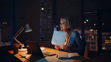 Geç kalmış patron sanal konferansta tartışıyor. Gece ofisindeki laptop web kamerasına evrakları gösteriyor. Sinirli iş kadını video görüşmesiyle çelişiyor. Gergin yönetici iş problemini tartışıyor 