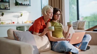 Modern oturma odasında bilgisayar arayan lezbiyen kızlar. Evcil kadınlar kanepeye sarılıp internetten kahve alıyorlar. Mutlu genç çift e-ticaret mağazasından bir şeyler sipariş ediyor. Dairede kucaklaşıyorlar.