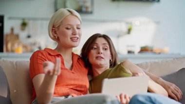 Modern evdeki rahatlamış LGBT çifti tablet bilgisayarı arıyor. Oturma odasındaki koltukta dijital cihaz kullanan sevimli insanlar. Mutlu kız arkadaşlar birlikte rahat bir evde vakit geçiriyorlar. 