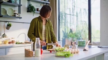 Yakın plan esmer, salata hazırlayan sabah penceresinin yanında yemek pişirme uygulaması. Odaklanmış kadın sebze doğruyor vejetaryen yemeği yapıyor. Bakan kadın, sağlıklı kahvaltı rutinine konsantre oluyor. 