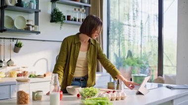 Odaklanmış esmer, kahvaltı hazırlıyor. Modern tablette video seyrediyor. Ciddi bir kadın evde besleyici yemek hazırlıyor. Mutfakta yemek fikirleri için ekranı kullanan konsantre bir kız.