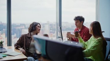 Ofis masasında kahve fincanı tutan gevşek çaylaklar konuşuyor. Üç iş arkadaşı modern iş yerinde gevşek sohbetin tadını çıkarırlar. Gülümseyen genç çalışanlar iş yerinde haberleri paylaşıyorlar. 