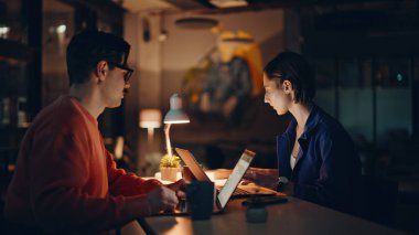 İş ortakları gece ofisinde lamba ışığında yakın plan çekimde çalışıyor. Ciddi bir profesyonel yönetici karanlık iş yerindeki kadın meslektaşına belge gösteriyor. Kapalı alanda evrak işleriyle uğraşan fazla mesai işçisi..