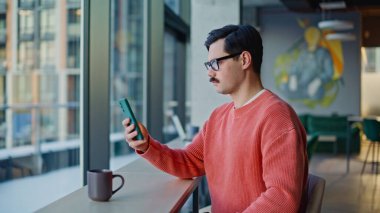 Modern kafeterya masasında oturan ciddi hippi görünümlü akıllı telefon. Genç bıyıklı adam şehir kafesinde cep telefonu uygulamasına bakıyor. Gözlüklü serbest çalışan cep telefonu bildirimi okuyor.