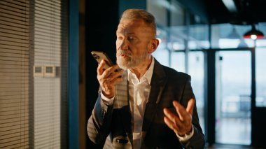 Günbatımı ışıklarıyla aydınlatılan akıllı telefon yürüyüş koridorunun ses notalarını dikte eden yönetici. Yaşlı işadamı ofis koridorunda sesli mesaj kaydediyor. Sakallı müdür hoparlörden konuşuyor.