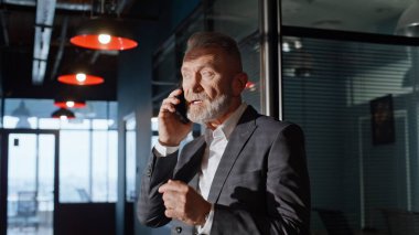 Sakallı yönetici akıllı telefon konuşuyor ofis koridorunda zarif bir takım elbise giyiyor. Olgun iş adamı partnerlerini cep telefonuyla arıyor. Yasli CEO cep telefonuyla is planini tartisiyor.