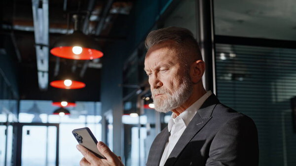 Старший предприниматель смотрит мобильный телефон в офисном коридоре крупным планом. Нахмуренный пожилой бизнесмен, читающий телефонные сообщения в холле. Серьезный владелец бизнеса отправляет электронную почту на смартфоне в помещении